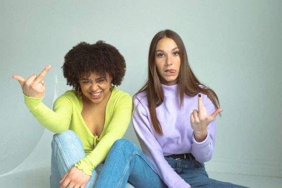 black woman with full afro in a bright yellow top flipping the middle finger white woman in light purple hoodie giving the bird to people who say when are you having kids