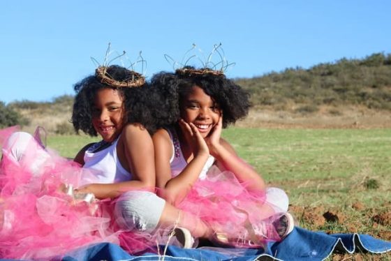 twin black girls, smileing in pink tutu's, on a green hillside, the twins are wearing brown twisted twig crowns, illustration best fertility now ivf twins article