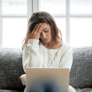 olive skin woman in a white jumper sat on a sofa loomking worried at a lap top screen, illustrating the Dear Future Egg Donor Mama artilce on best fertility now